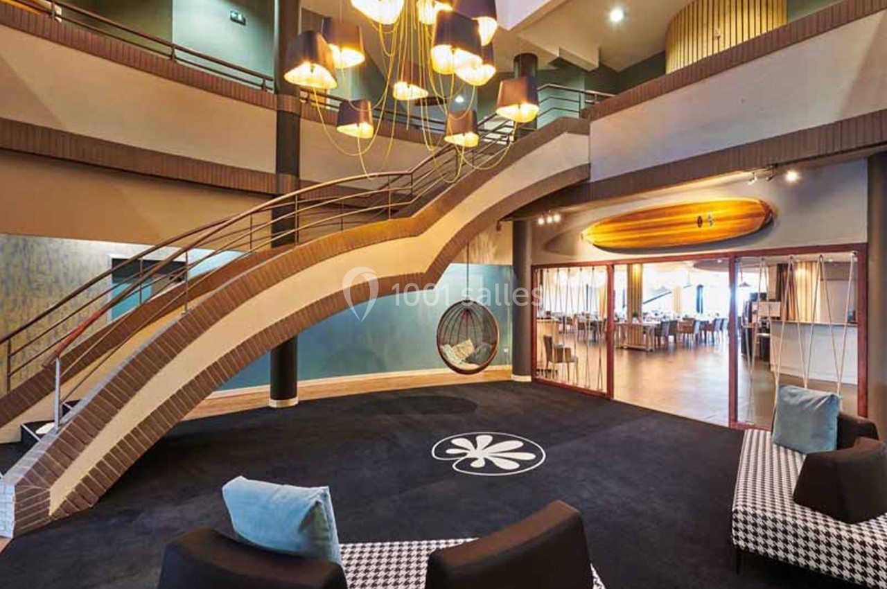 Hall d'entrée avec escalier courbe en briques, grand lustre à abat-jours, fauteuils en velours noir et coussins bleus, et une salle de restaurant visible à droite.