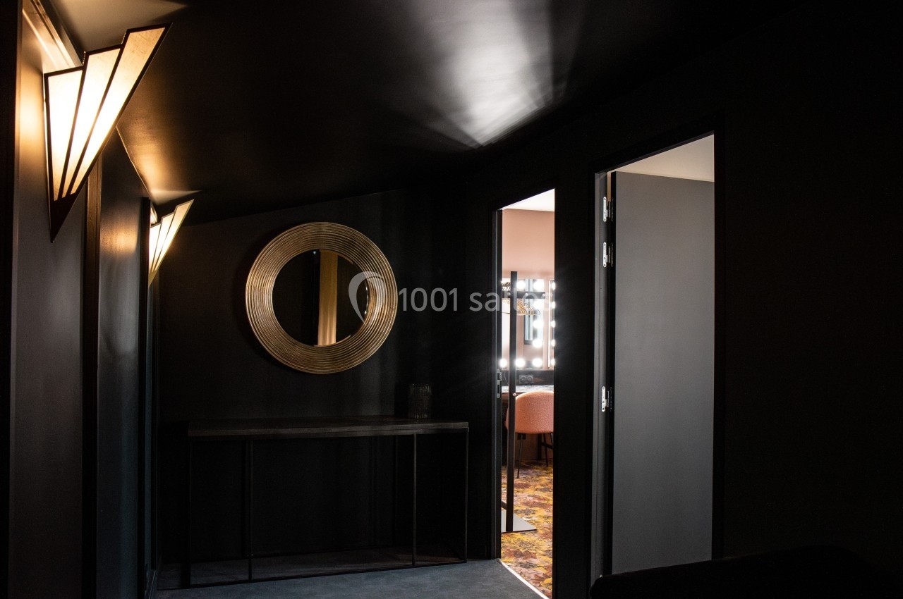 Couloir sombre avec un miroir rond doré, une console noire et des portes ouvertes laissant entrevoir des pièces éclairées.