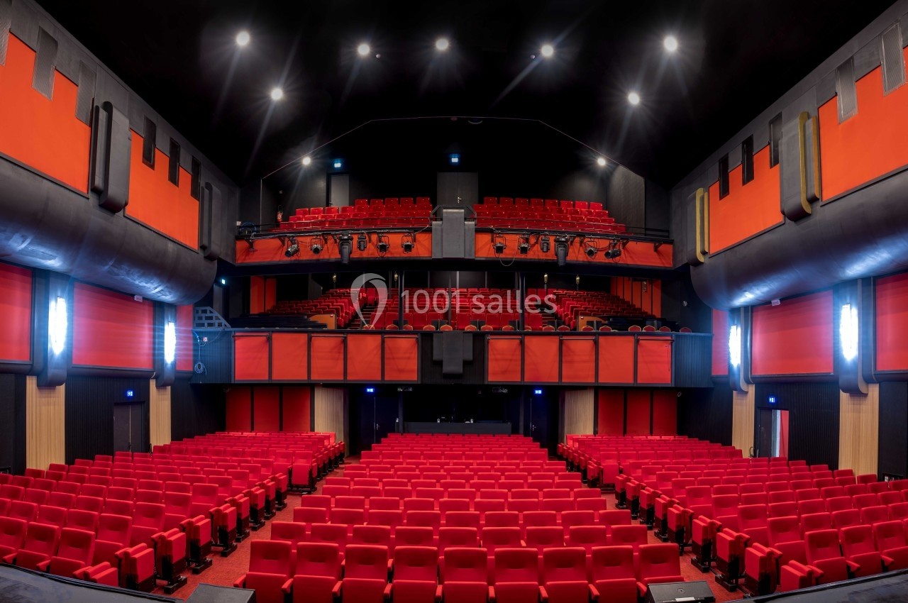 Vue intérieure d'une salle de théâtre moderne avec des rangées de sièges rouges et une scène au fond.
