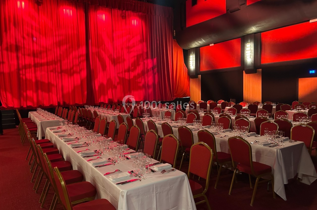 Salle de réception avec des tables dressées, nappes blanches et chaises rouges, devant une scène avec rideaux rouges.