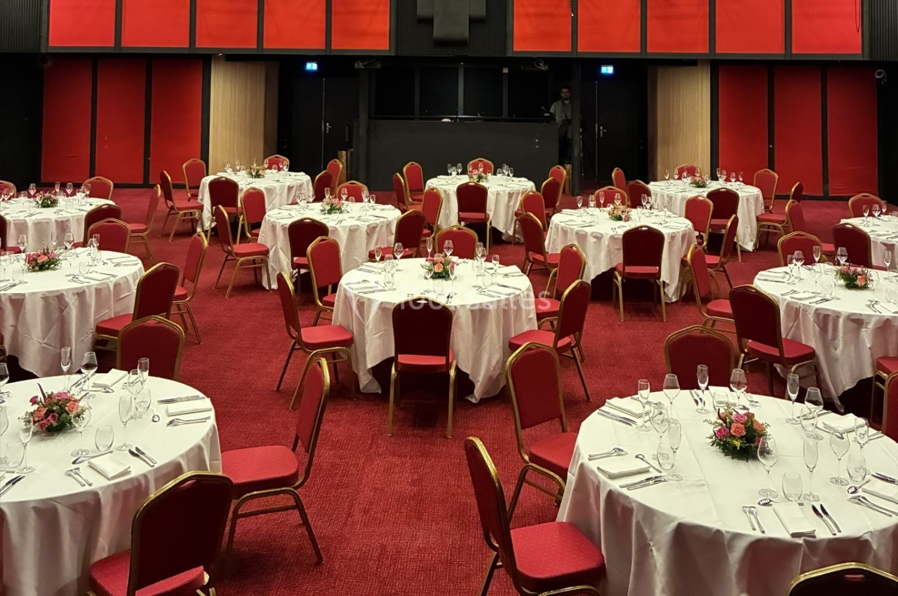 Salle de réception aménagée avec des tables rondes dressées, nappes blanches, chaises rouges et décorations florales.