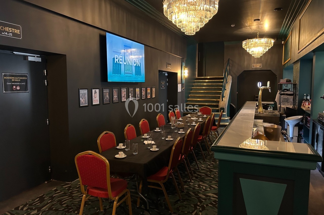 Salle de réunion avec table dressée, chaises rouges, bar à droite, écran mural et lustres suspendus.
