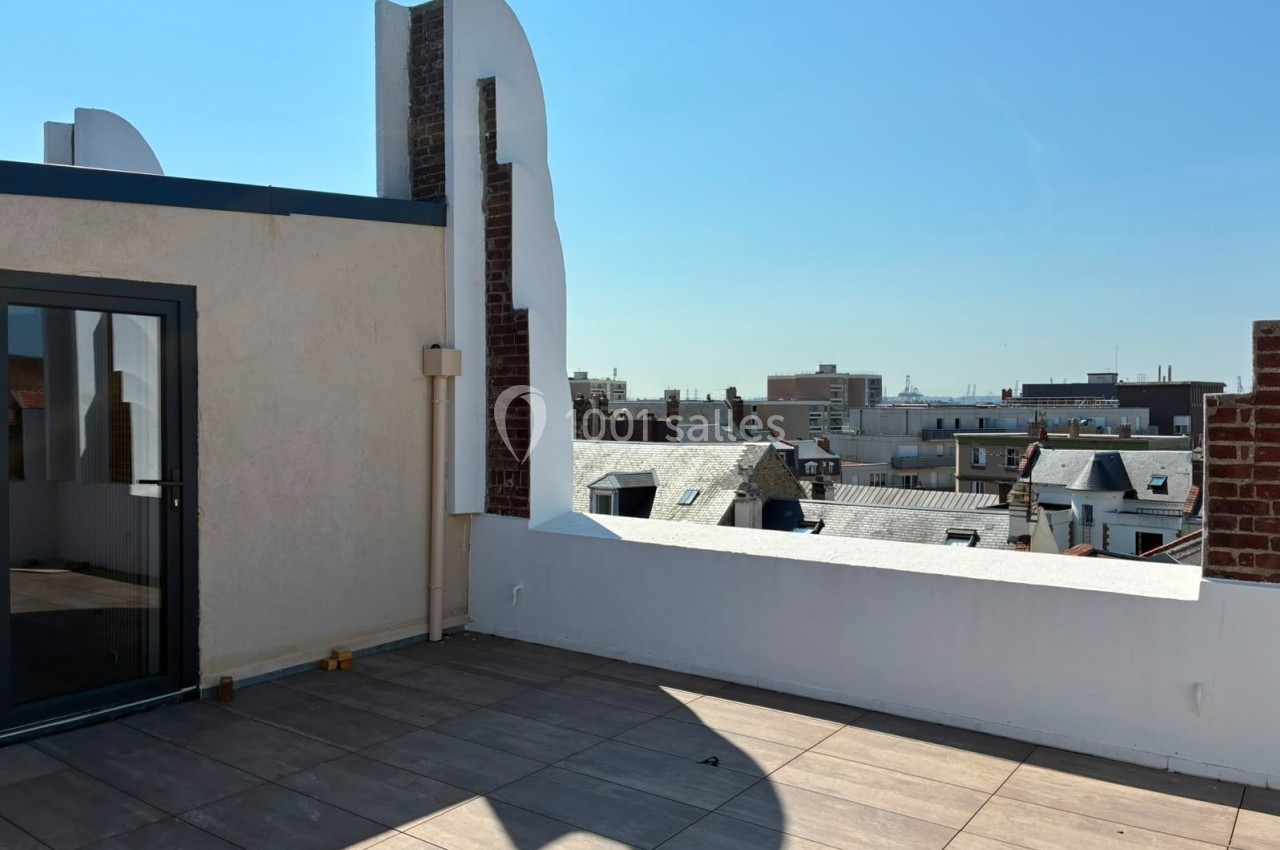 Terrasse ensoleillée avec sol en carrelage, entourée de murs blancs et vue sur les toits d'une ville.