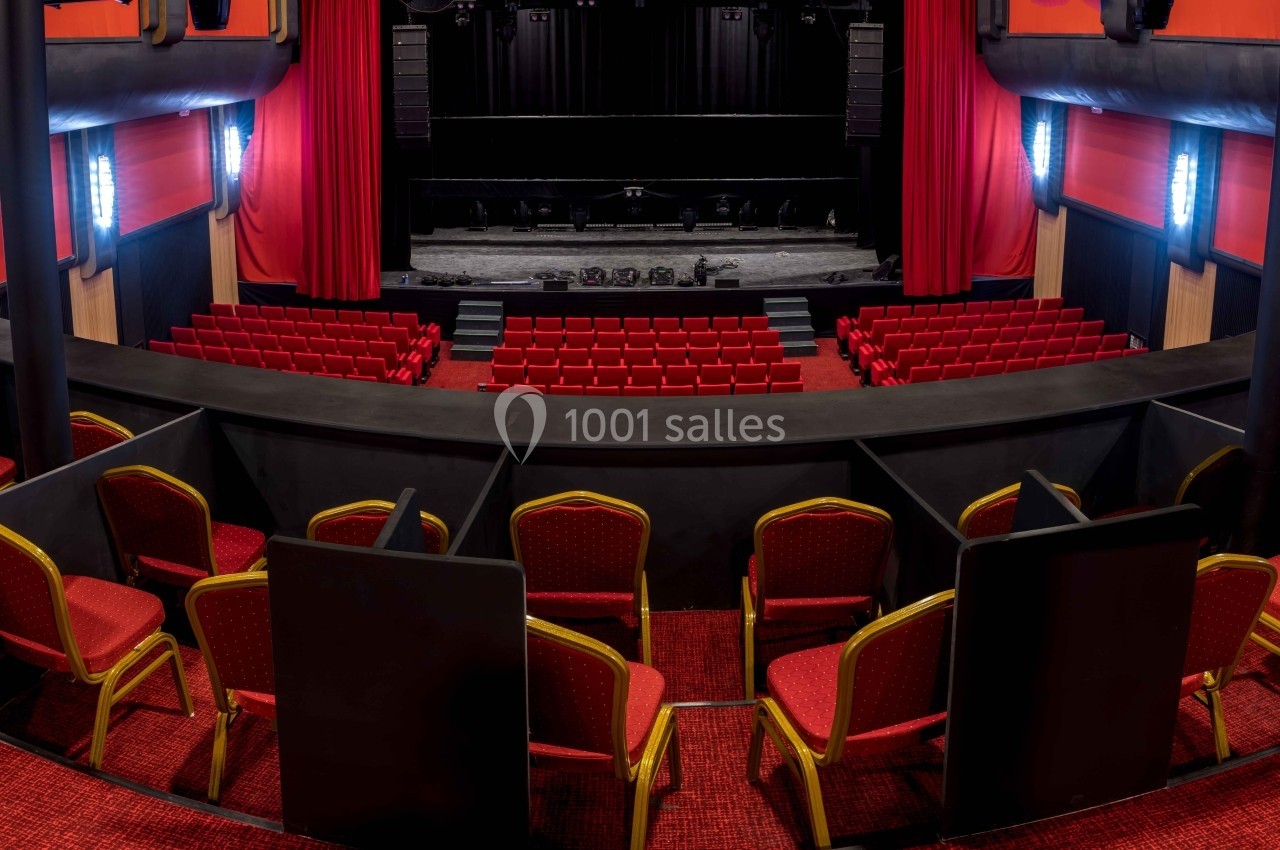 Vue intérieure d'un théâtre avec des rangées de sièges rouges, une scène centrale et des rideaux rouges sur les côtés.