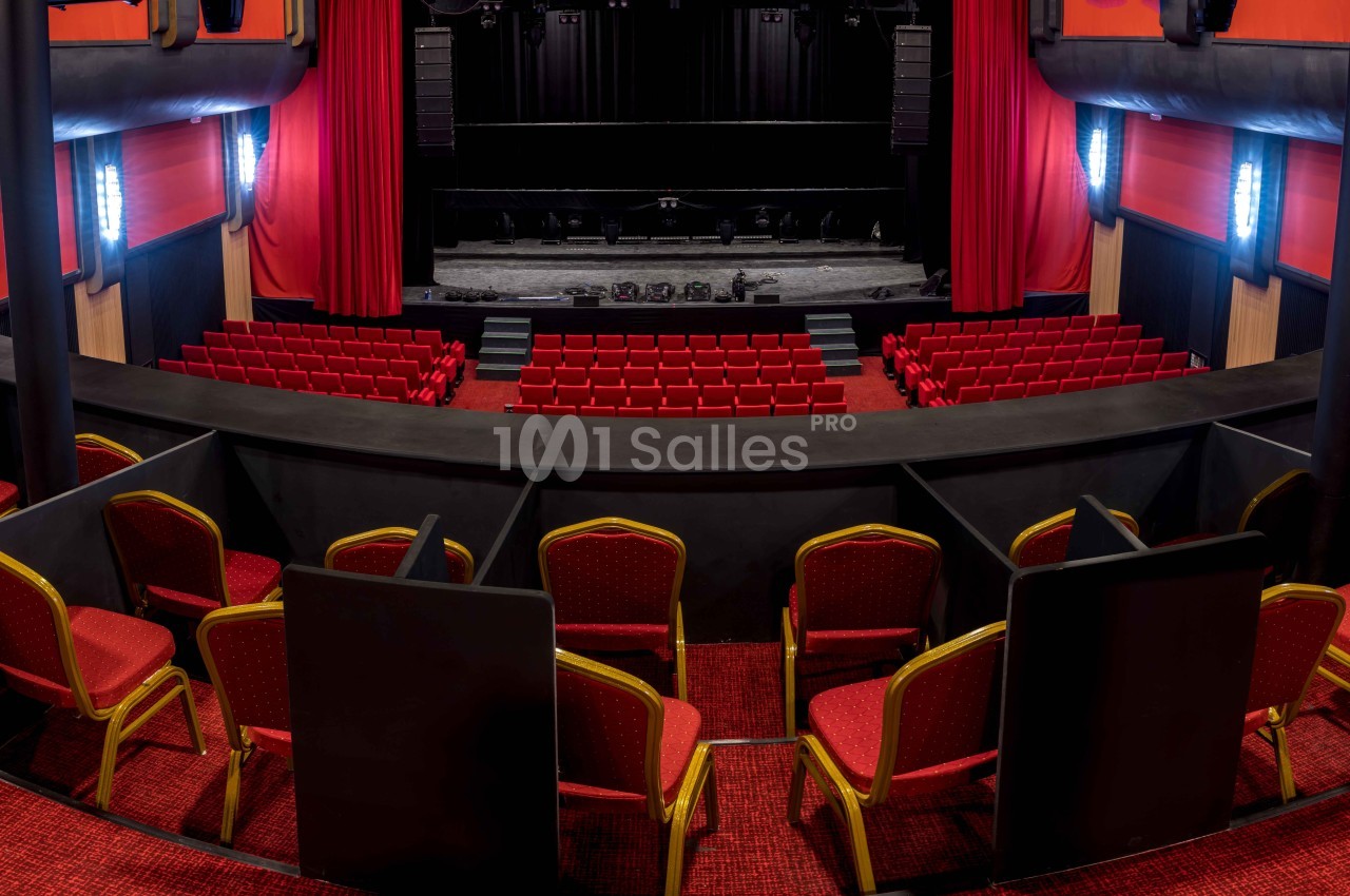 Vue intérieure d'un théâtre avec des rangées de sièges rouges, une scène centrale et des rideaux rouges sur les côtés.