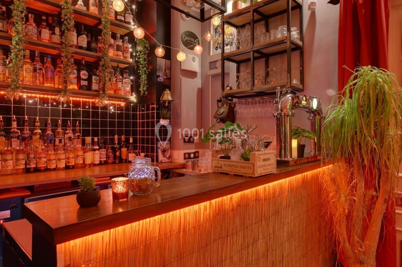 Bar chaleureux avec comptoir en bois, étagères remplies de bouteilles, lumières tamisées et décorations végétales.