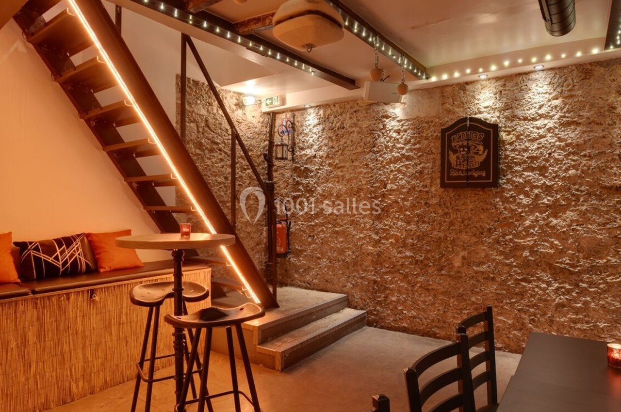 Espace intérieur chaleureux avec murs en pierre, escalier éclairé, table haute et banquette avec coussins colorés.