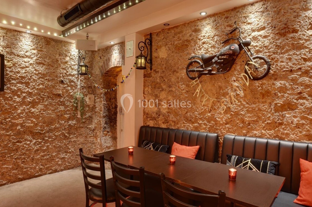 Salle décorée avec des murs en pierre, une moto accrochée, des lanternes murales et une table entourée de chaises.