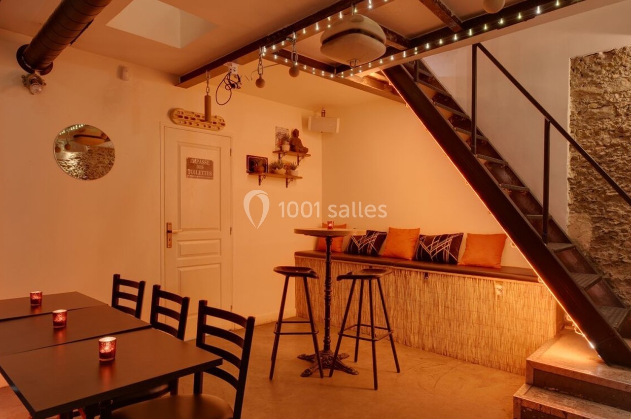 Salle chaleureuse avec tables, chaises noires, banquette orange, escalier métallique et murs en pierre apparente.