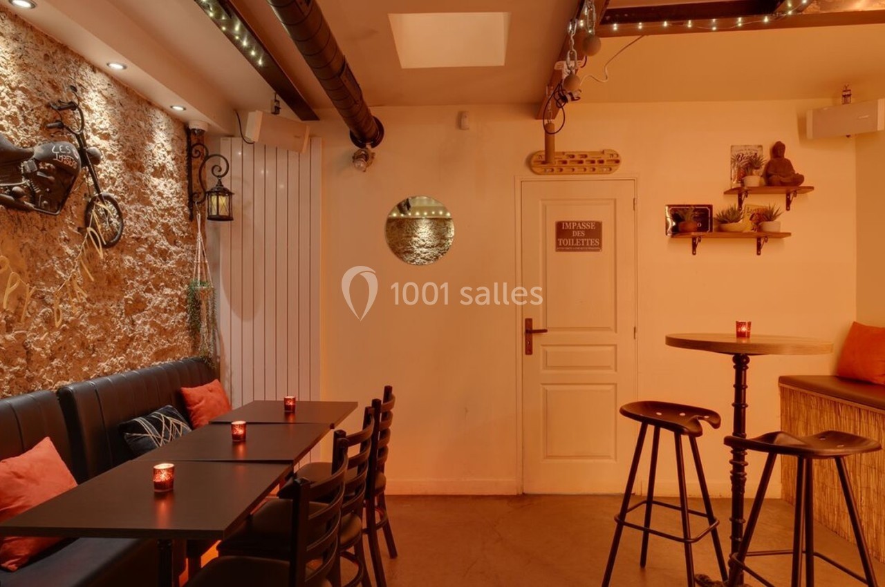 Salle chaleureuse avec murs en pierre, tables hautes, banquette noire, coussins orange et éclairage tamisé.