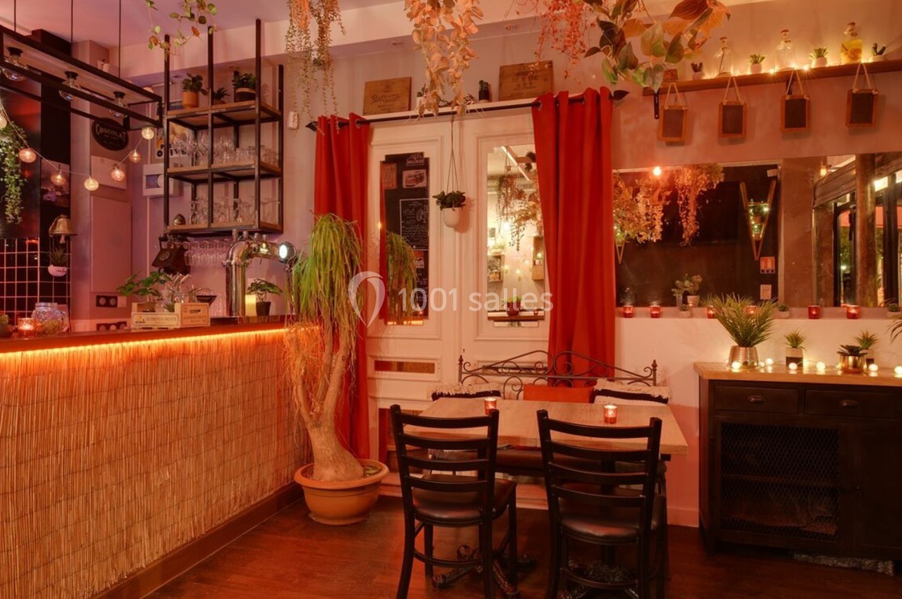 Intérieur d'un bar cosy avec éclairage tamisé, plantes suspendues, comptoir en bois et petite table entourée de chaises.