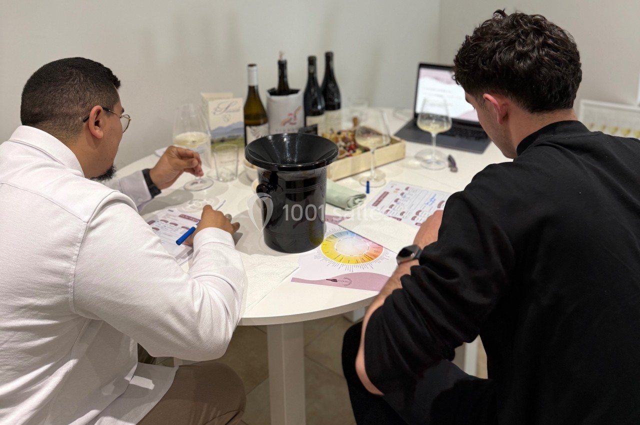 Deux hommes assis à une table dégustent du vin tout en prenant des notes, avec des bouteilles et un ordinateur en arrière…
