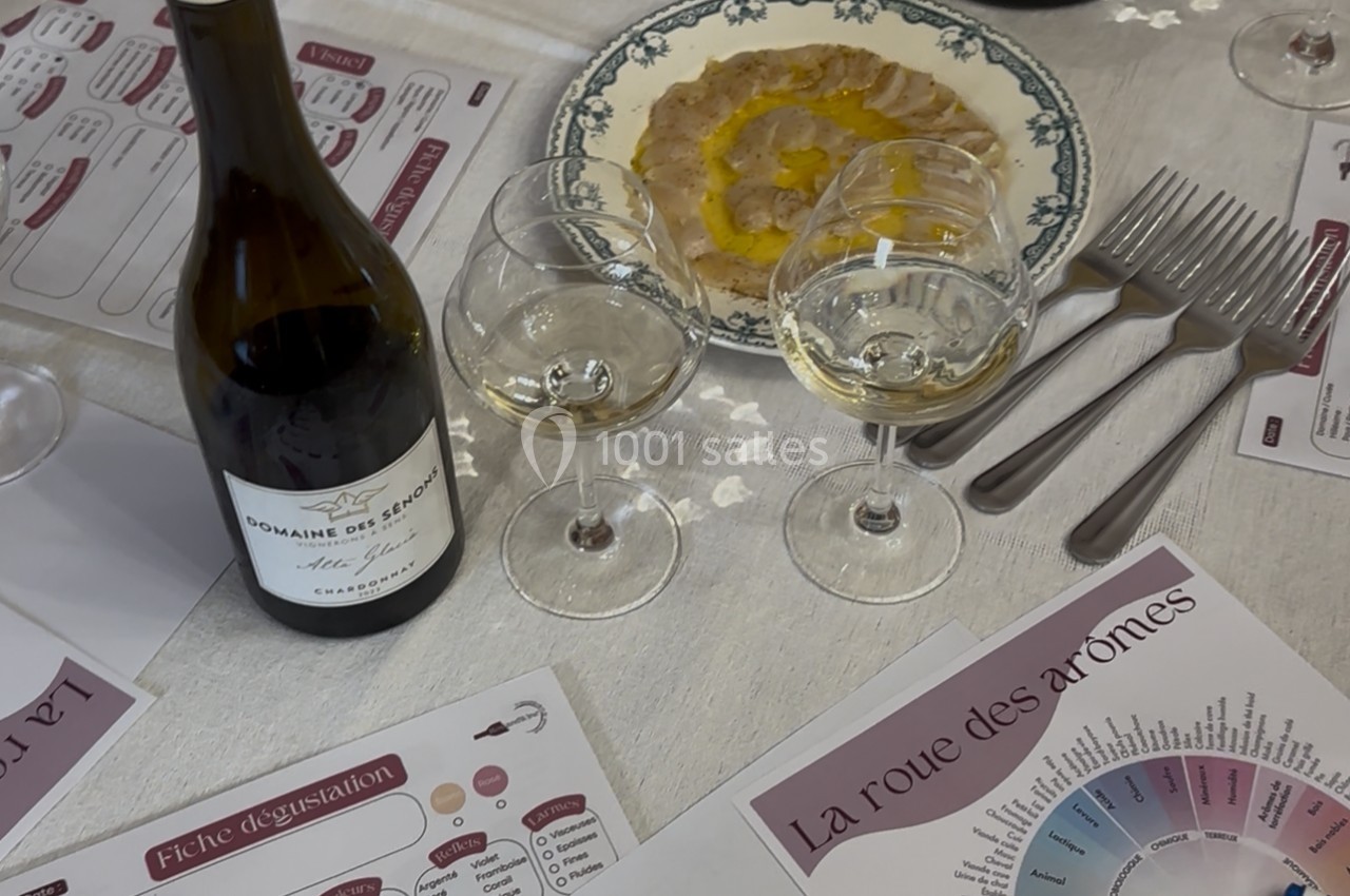 Bouteille de vin blanc, verres, assiette de carpaccio et documents de dégustation sur une table dressée.