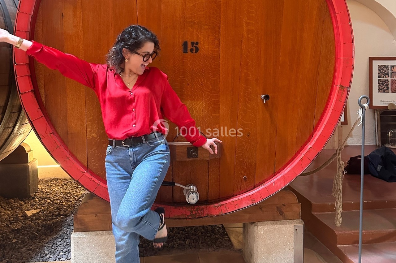 Femme en chemise rouge posant devant un grand tonneau en bois dans un environnement de cave à vin.