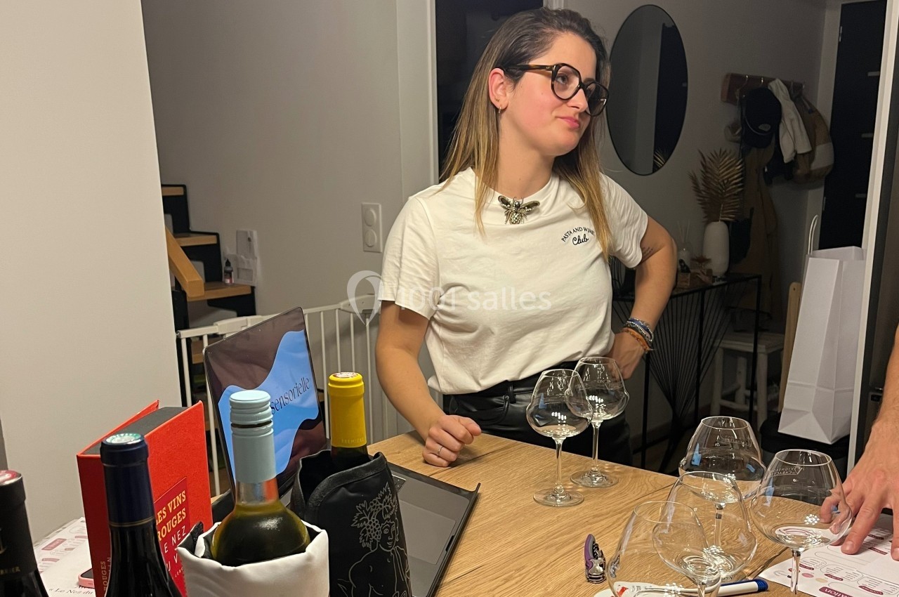 Une femme debout près d'une table avec des verres à vin, dans un intérieur moderne et éclairé.
