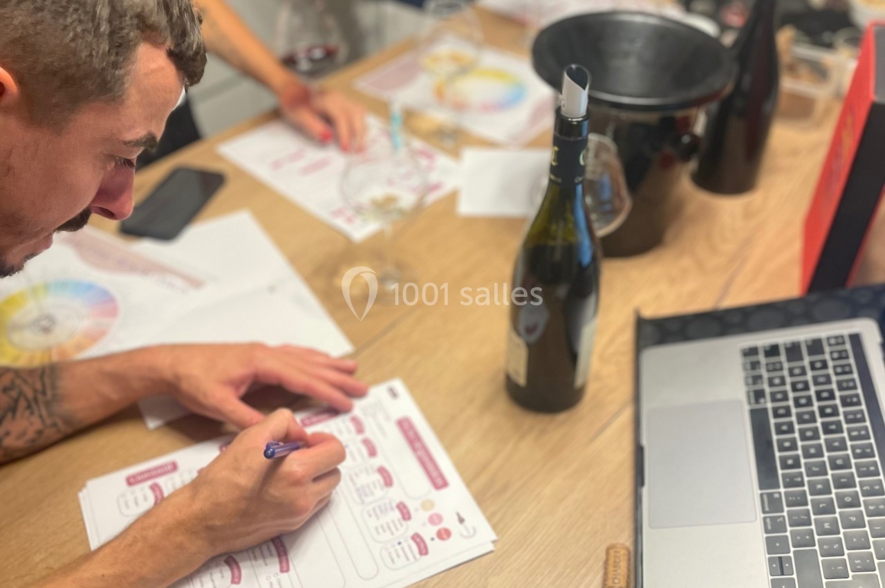Un homme remplit un formulaire de dégustation de vin sur une table avec une bouteille, un ordinateur et des documents.