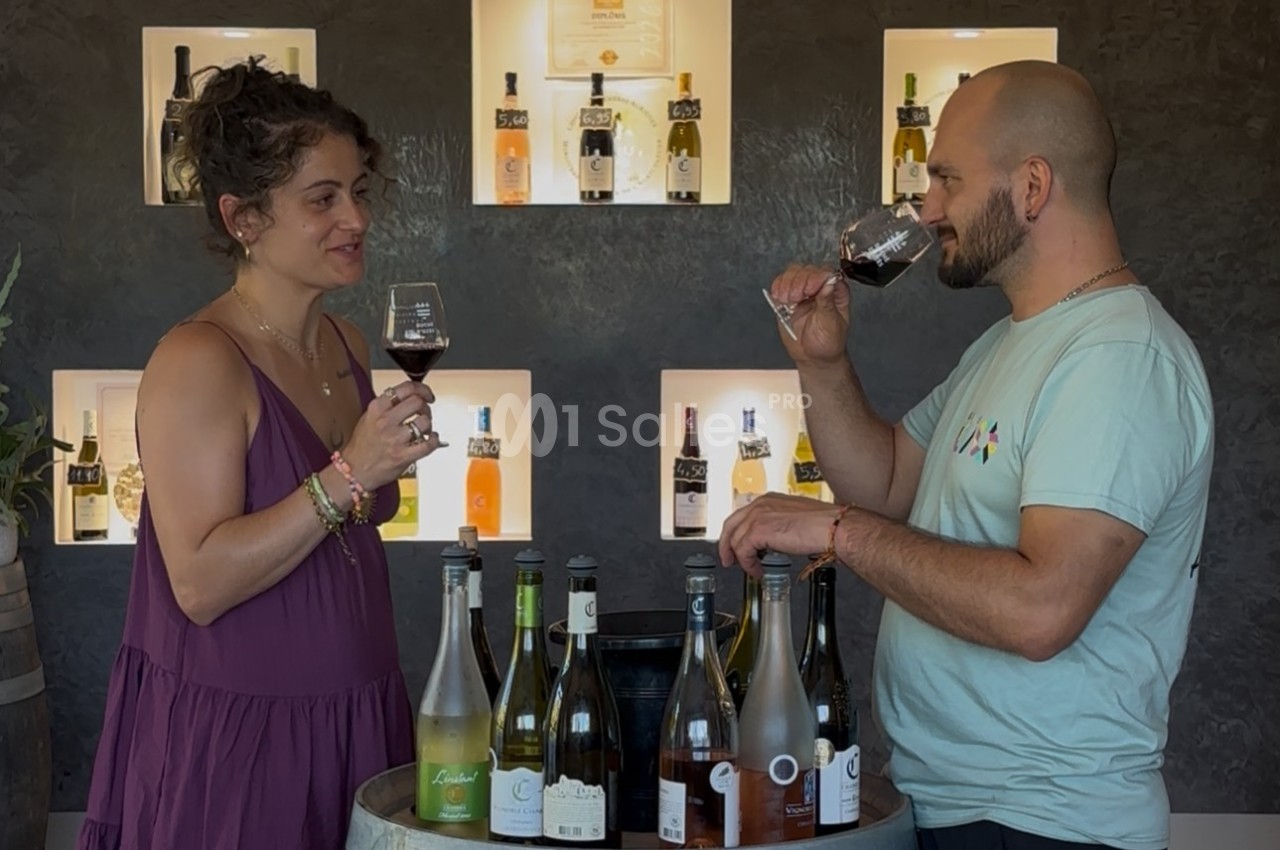 Deux personnes dégustent du vin devant un présentoir de bouteilles éclairé, avec un tonneau en premier plan.