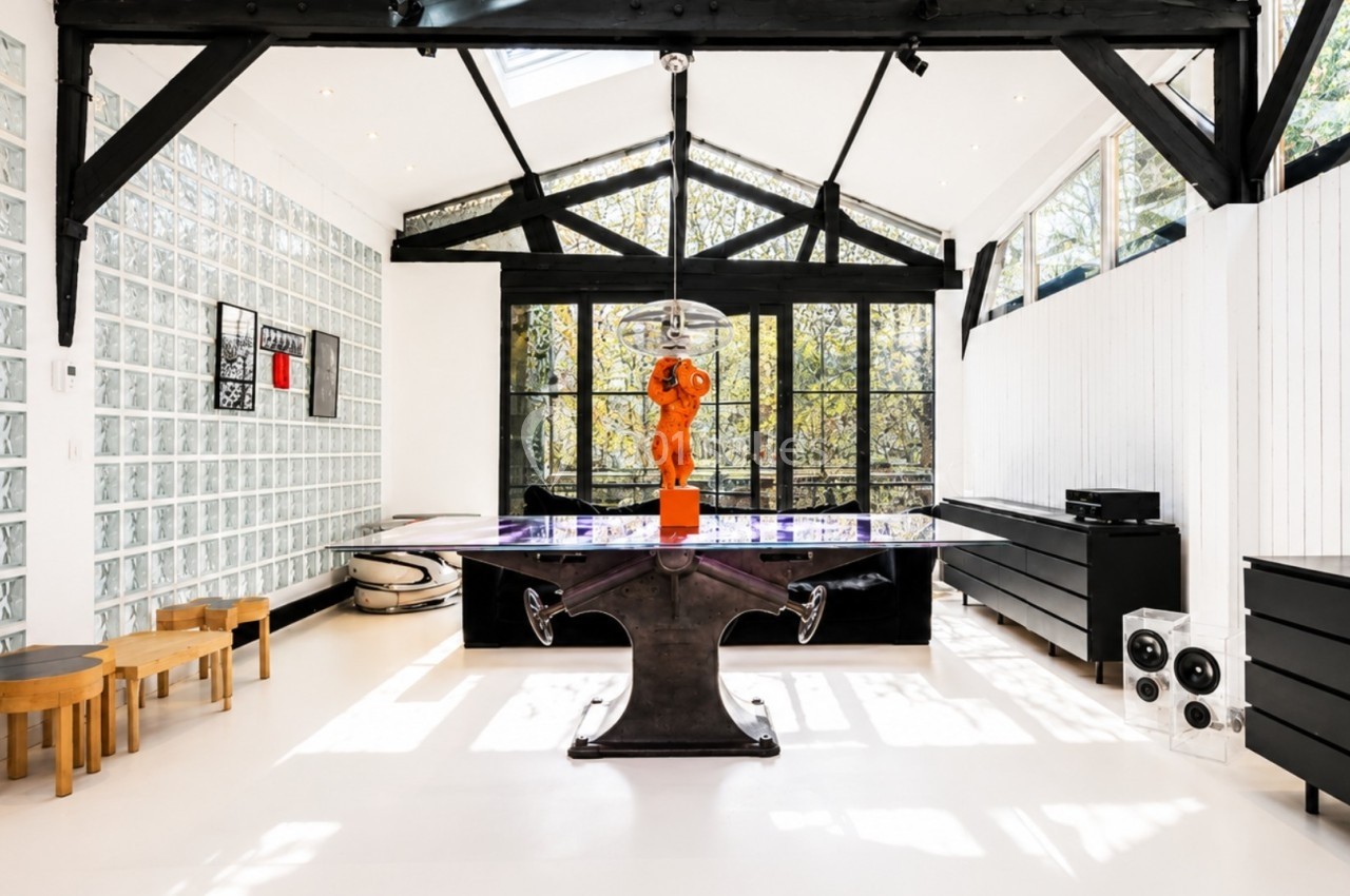 Salle lumineuse avec grande table centrale, murs vitrés, poutres noires apparentes et mobilier moderne.