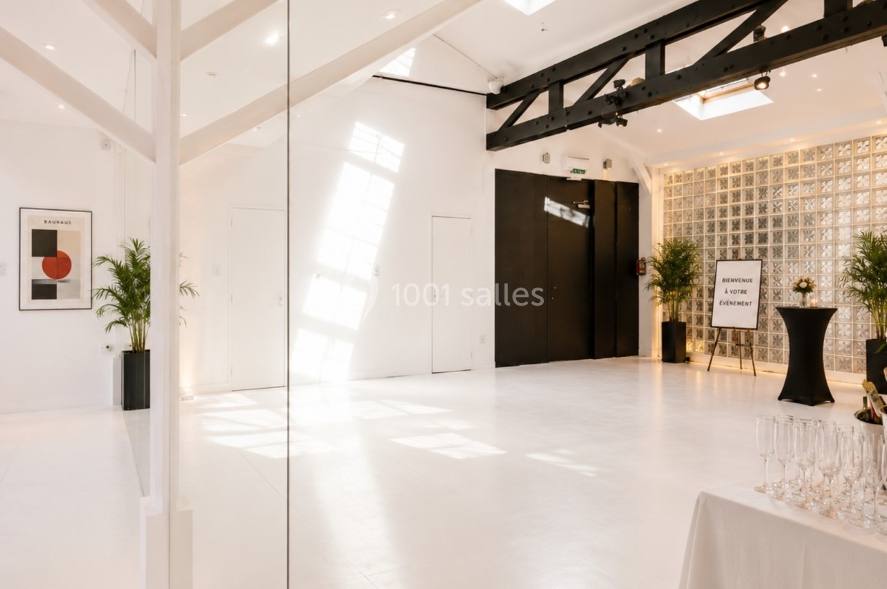 Salle lumineuse avec murs blancs, verrière, poutres noires apparentes, plantes décoratives et table avec flûtes de champagne.