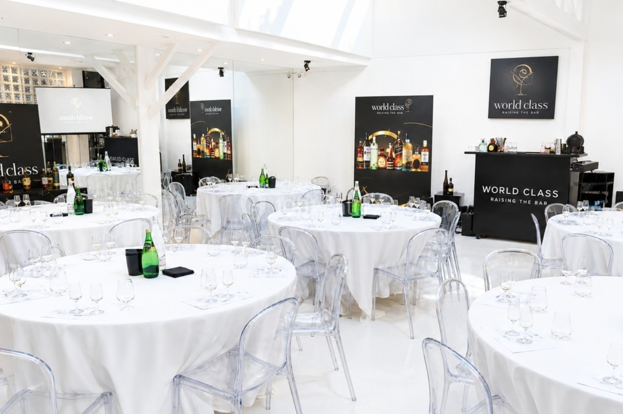 Salle lumineuse avec des tables rondes dressées, des chaises transparentes et des affiches ’World Class’ en arrière-plan.