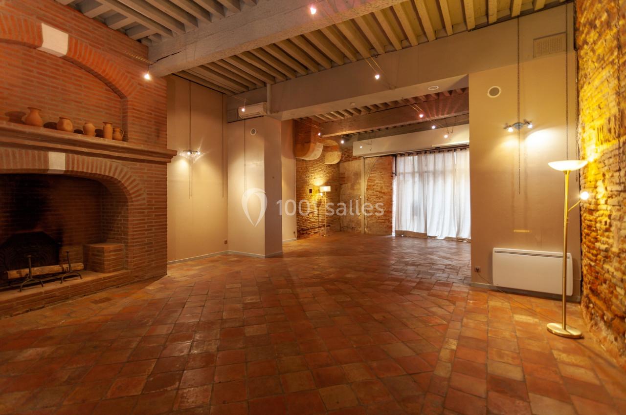 Salle vide avec sol en terre cuite, murs en briques, grande cheminée et lumière naturelle entrant par des rideaux.