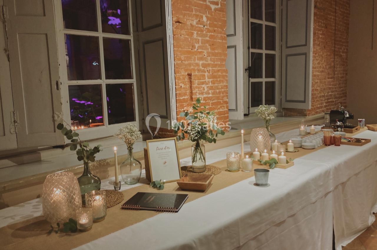 Table décorée avec bougies, fleurs et cadres, placée devant un mur en briques et des fenêtres ouvertes.