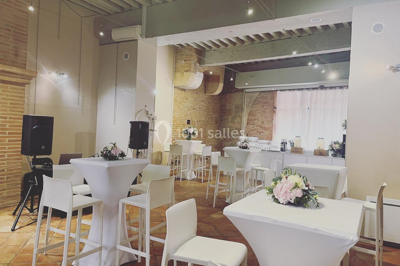 Salle lumineuse avec tables hautes blanches, chaises assorties, décorations florales et murs en briques apparentes.