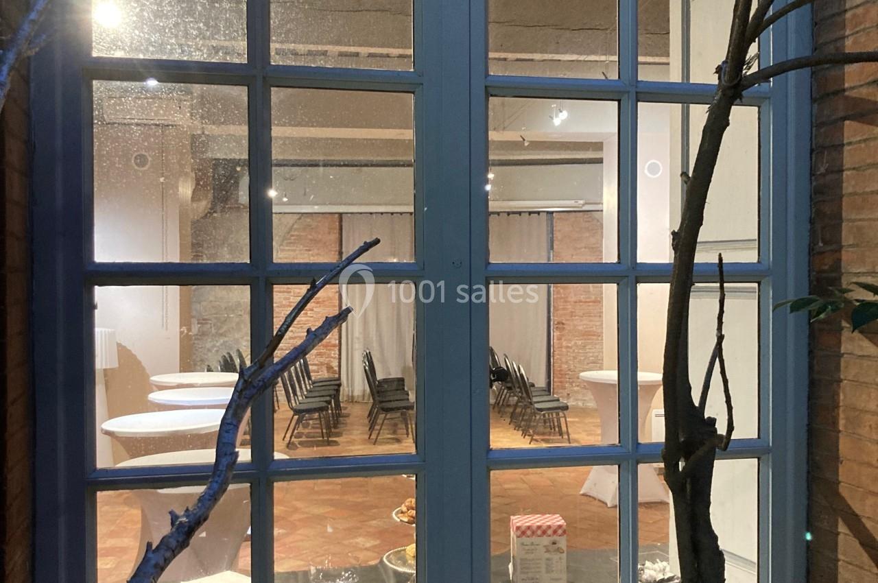 Vue à travers une fenêtre sur une salle éclairée avec des tables hautes, des chaises noires et un sol en terre cuite.