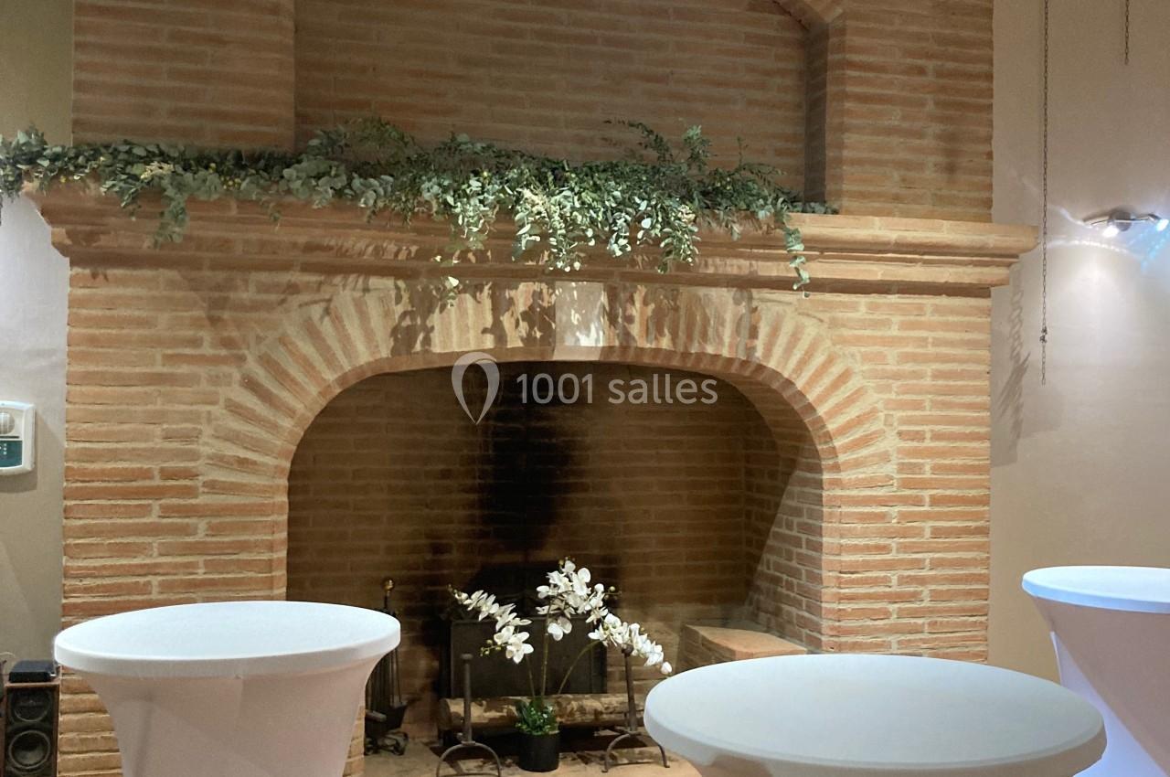 Cheminée en briques décorée de plantes, entourée de tables hautes drapées de blanc dans une salle au sol en terre cuite.