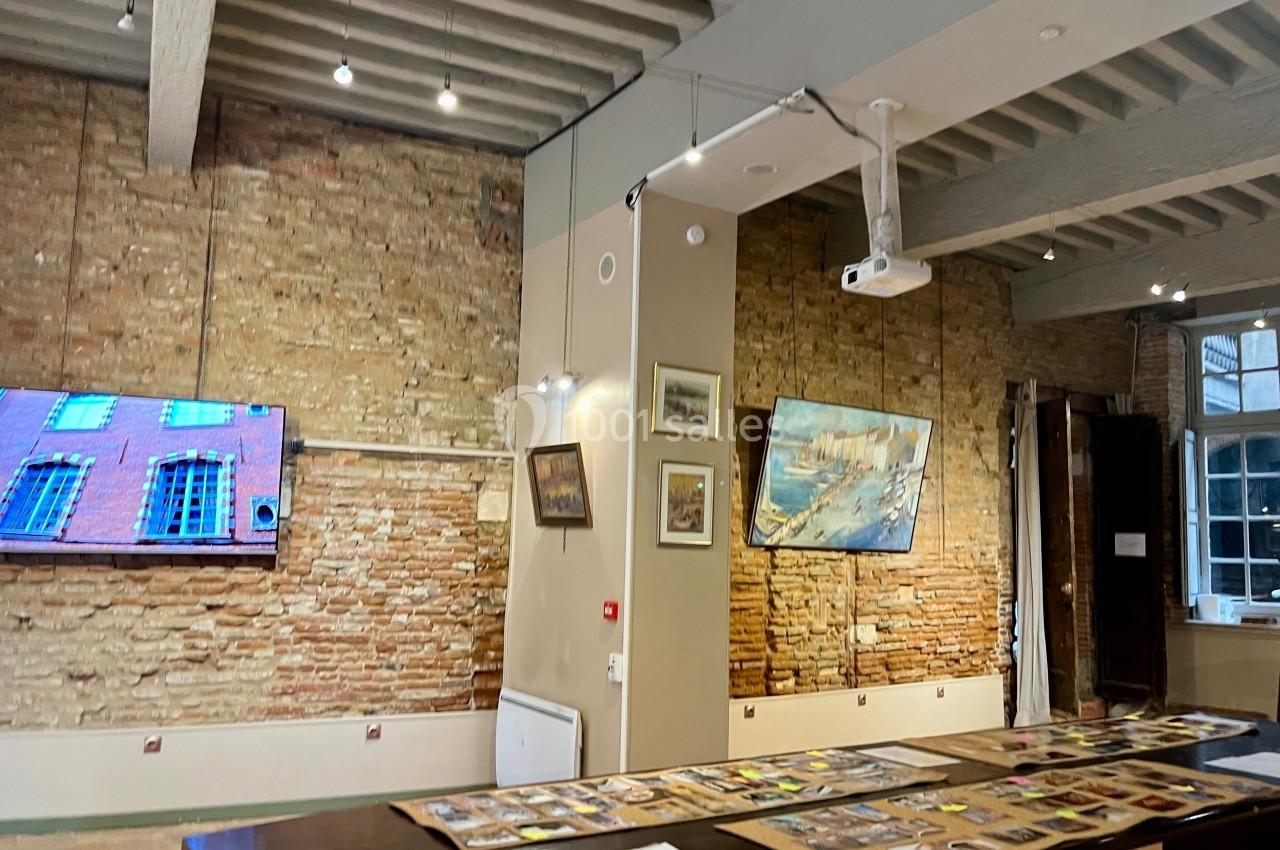 Salle d'exposition avec murs en briques, tableaux accrochés, écran mural et table présentant des documents.