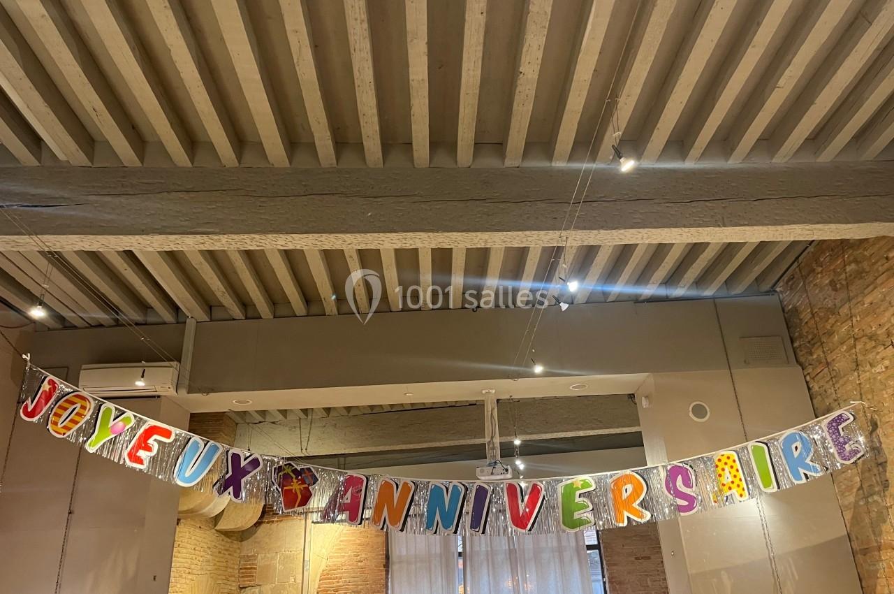 Guirlande colorée avec l'inscription ’Joyeux anniversaire’ suspendue dans une pièce au plafond en poutres apparentes.