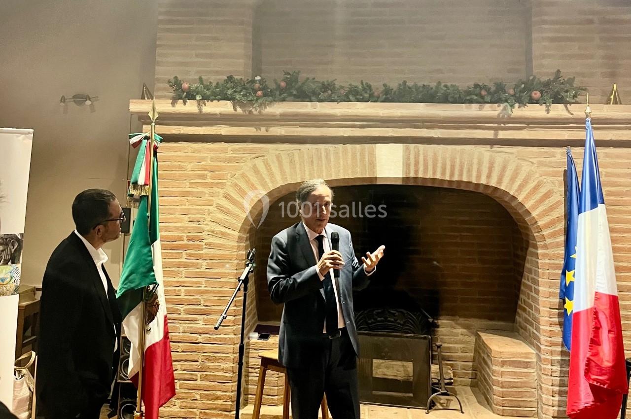 Un homme en costume parle au micro devant une cheminée en briques, près des drapeaux français et mexicain.