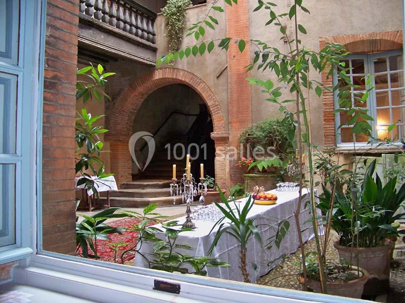 Cour intérieure avec table dressée, chandeliers, verres et fruits, entourée de plantes et d'arches en briques.