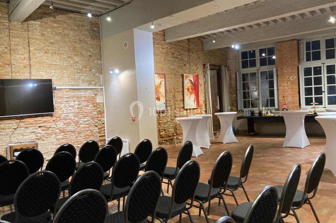 Salle avec des chaises alignées, tables hautes et buffet, murs en briques apparentes et grande fenêtre.