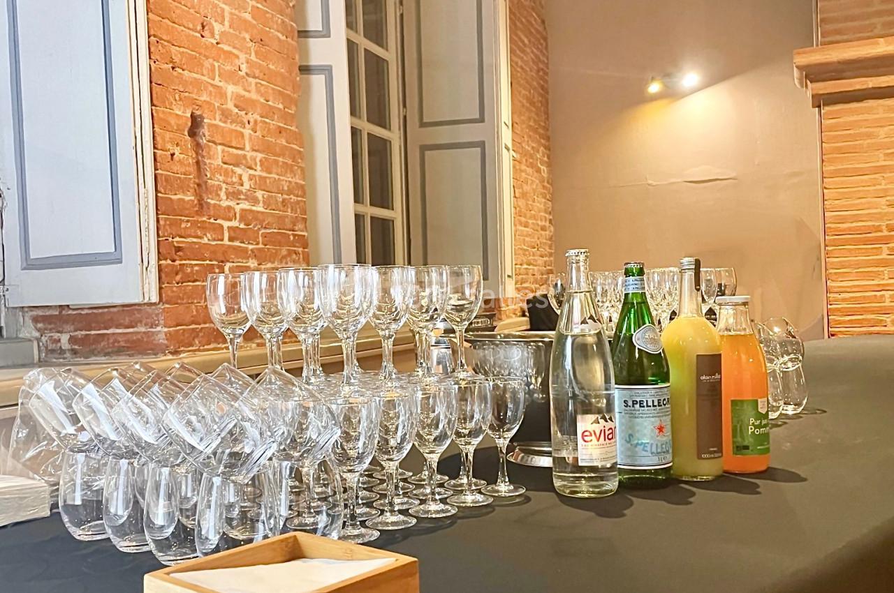 Table avec verres empilés, bouteilles d'eau et de jus, dans une pièce aux murs en briques et grandes fenêtres.