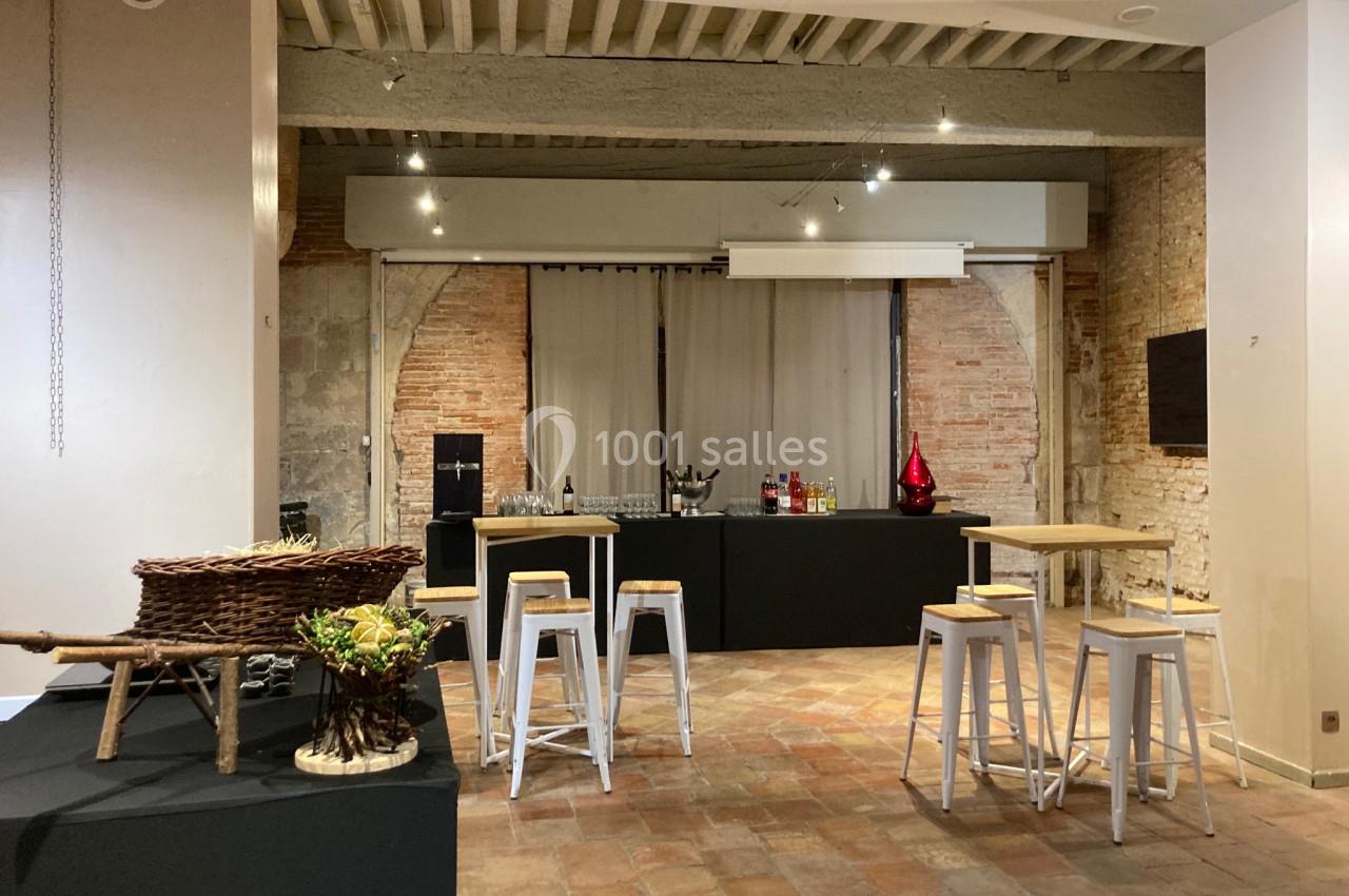 Salle avec murs en briques, tables hautes, tabourets blancs et un buffet équipé de boissons et décorations.