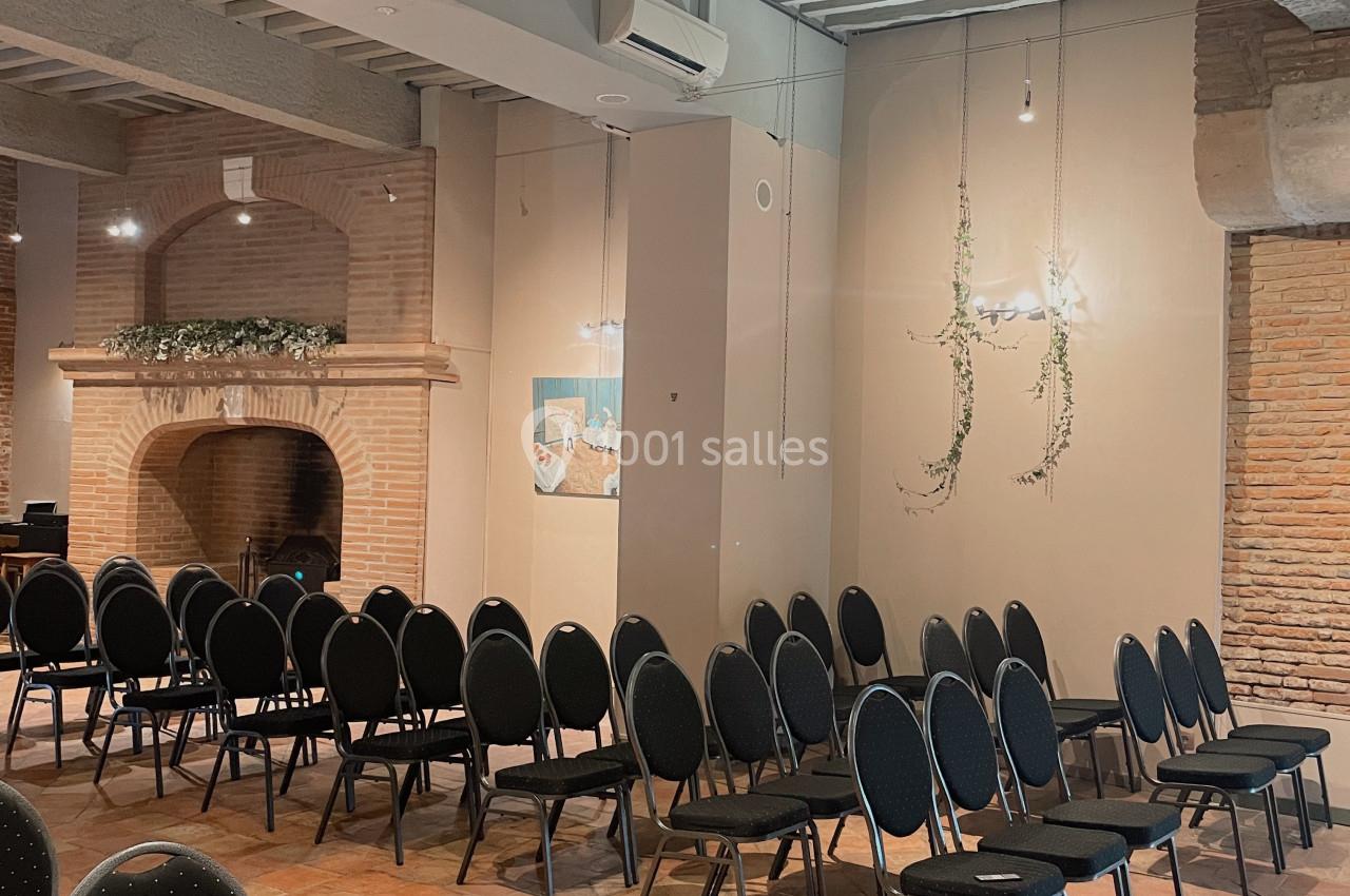 Salle avec des rangées de chaises noires alignées, sol en terre cuite, murs en briques et plafond avec poutres apparentes.