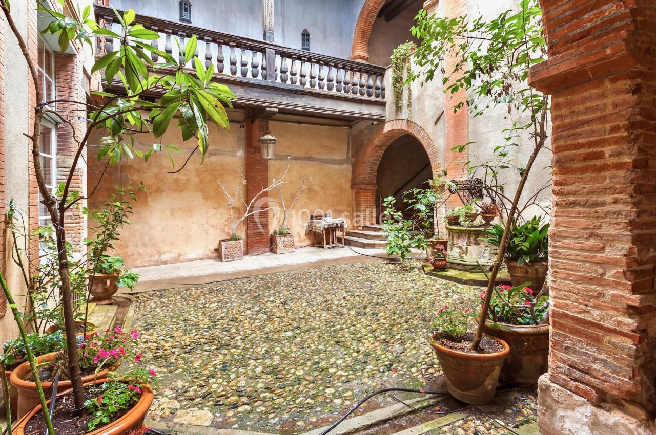 Cour intérieure pavée avec des plantes en pots, des murs en briques et un balcon en bois au-dessus.