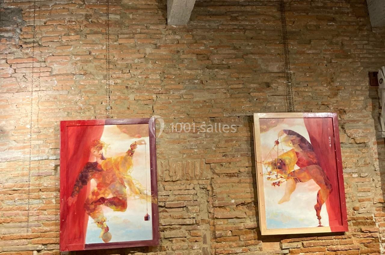 Deux tableaux colorés accrochés sur un mur en briques, avec des tables hautes blanches au premier plan.