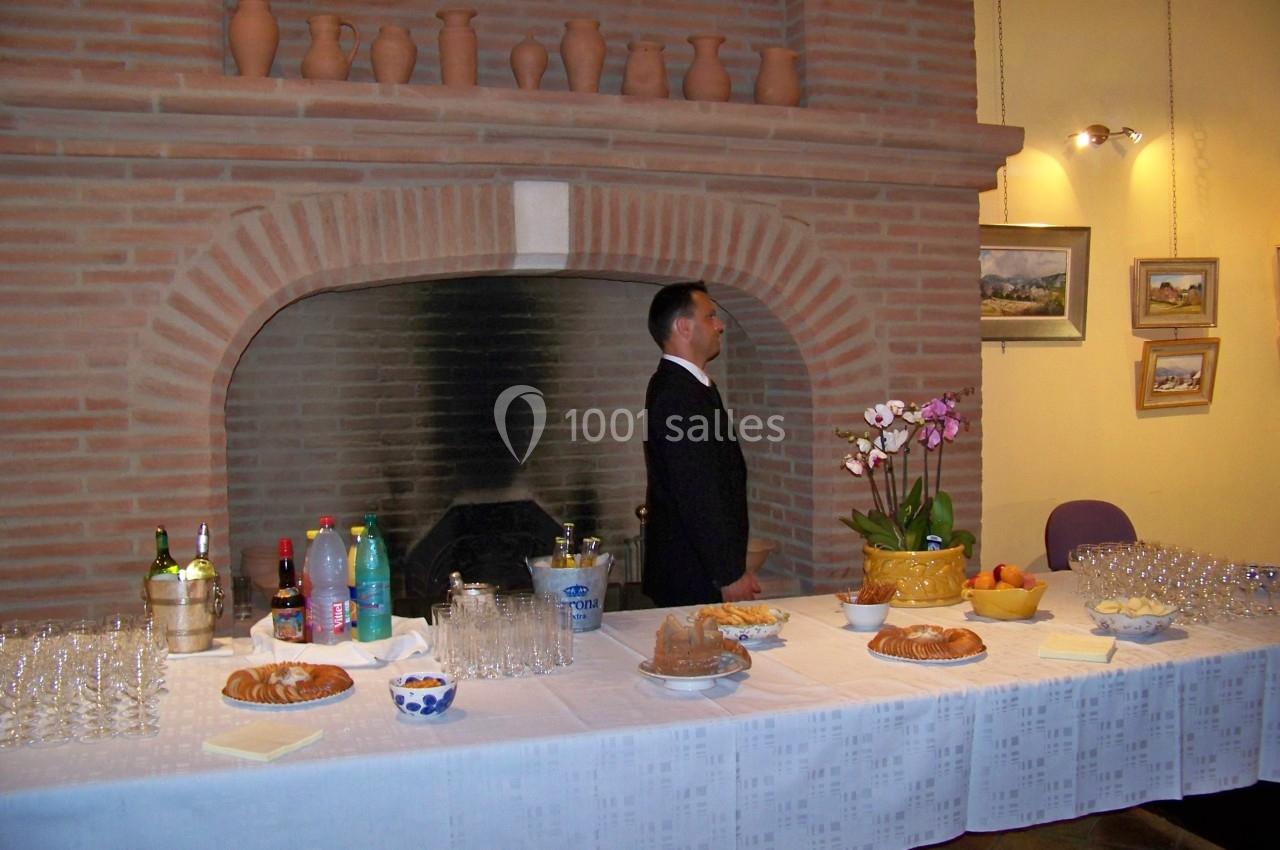 Table dressée avec boissons, pâtisseries, fruits et fleurs devant une cheminée en briques, un homme debout à côté.