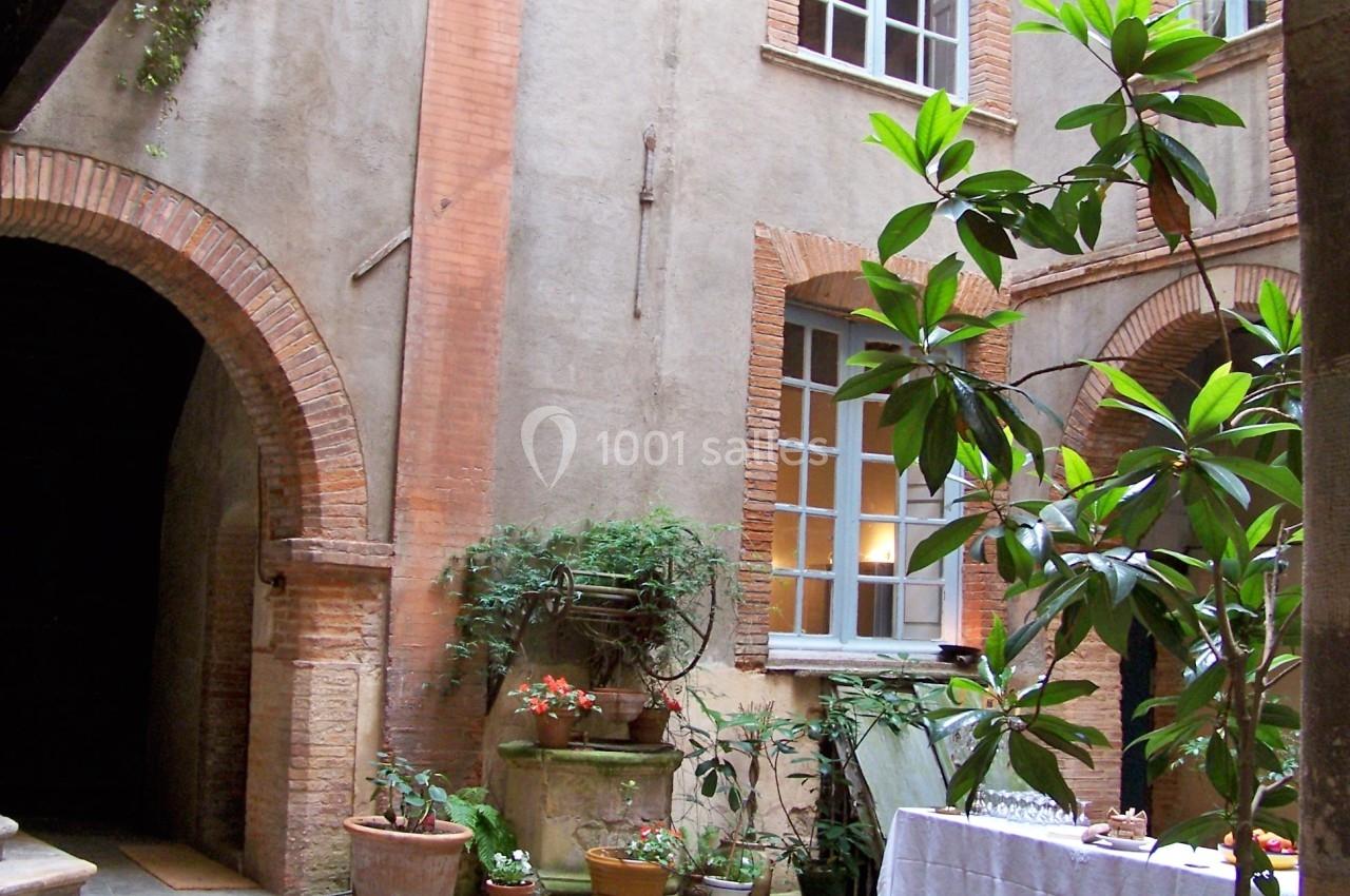 Cour intérieure avec murs en briques, tapis rouge, plantes en pot et table dressée sous une lumière naturelle.