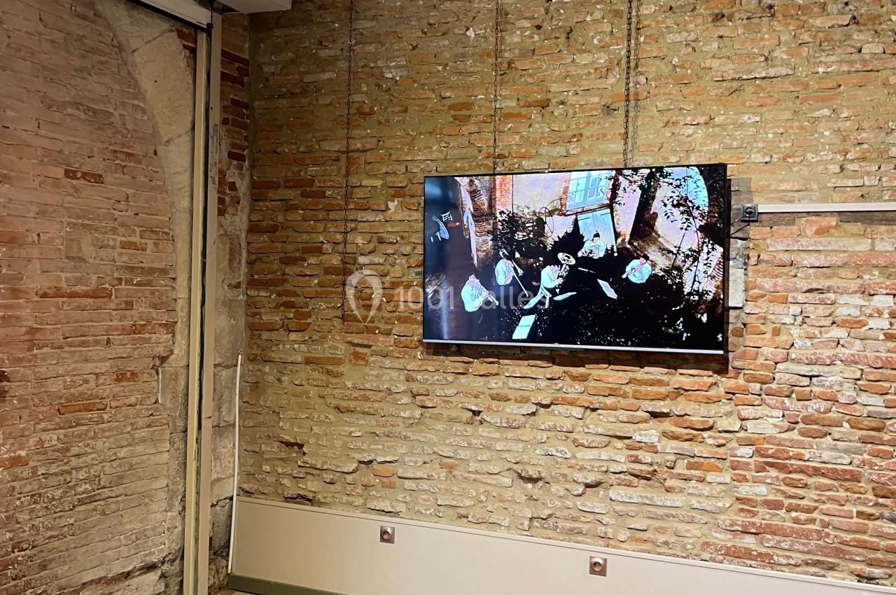 Mur en briques apparentes avec un écran fixé affichant une vidéo, sol en carrelage et plafond avec poutres en bois.