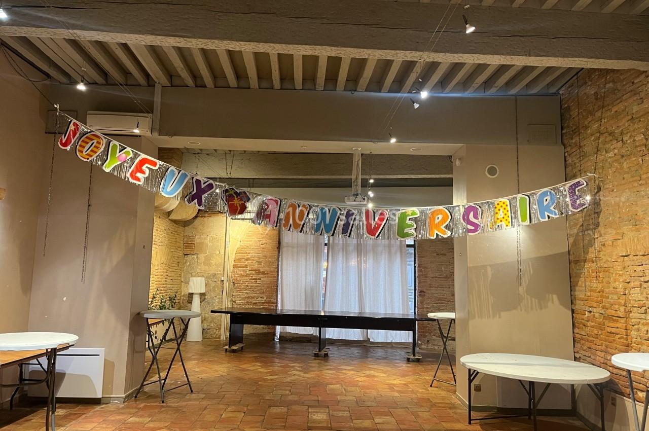 Guirlande colorée ’Joyeux Anniversaire’ suspendue dans une salle avec murs en briques et tables rondes vides.