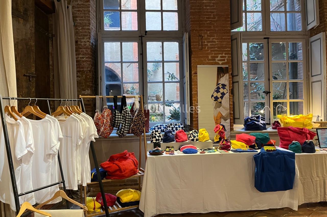 Présentoirs avec vêtements blancs sur cintres et accessoires colorés exposés sur des tables dans une pièce lumineuse.