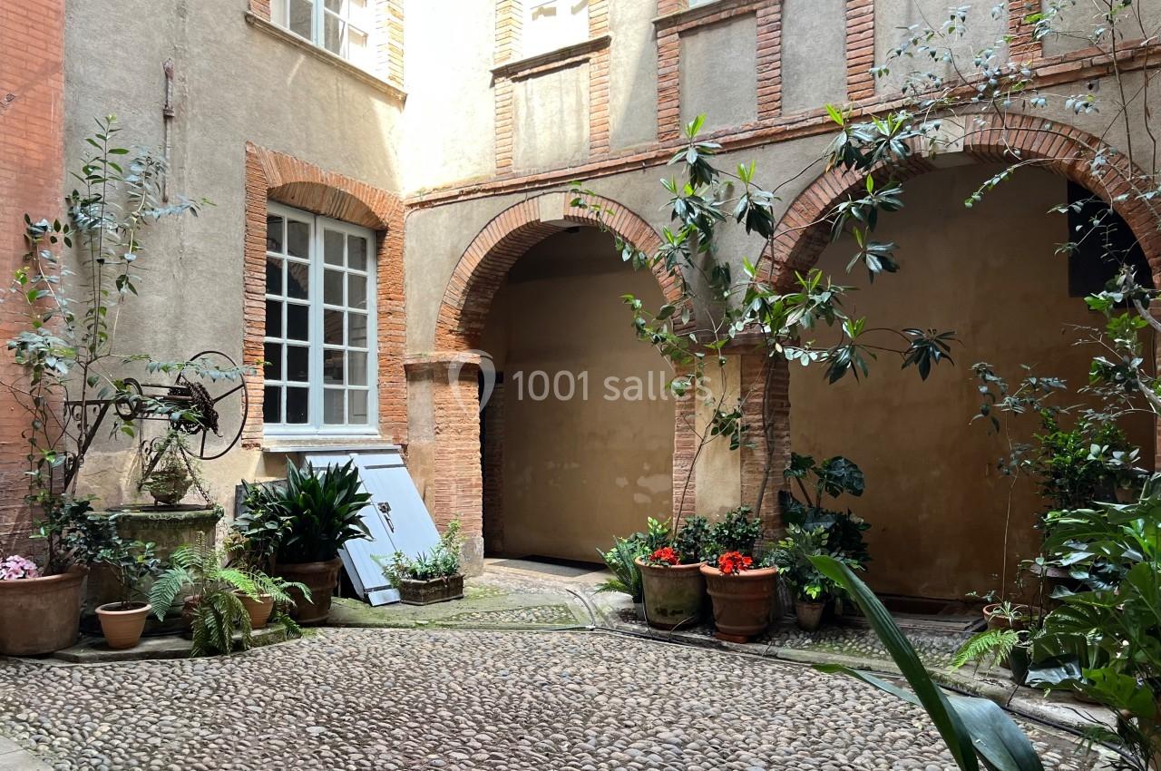 Cour intérieure avec pavés, plantes en pots, arches en briques et mur beige, éclairée par une lumière naturelle.