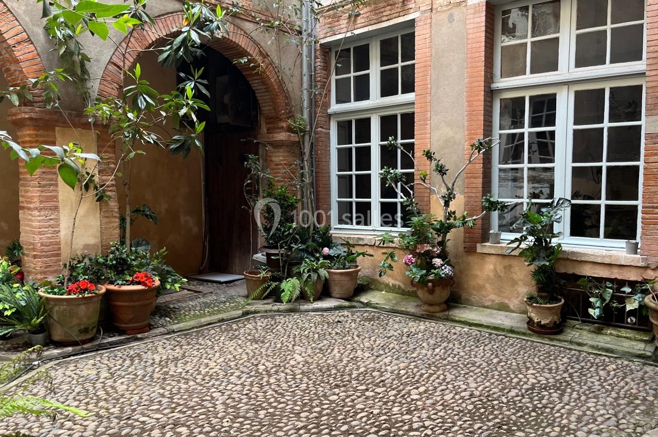 Cour intérieure pavée avec des plantes en pots, entourée de murs en briques et fenêtres à carreaux.