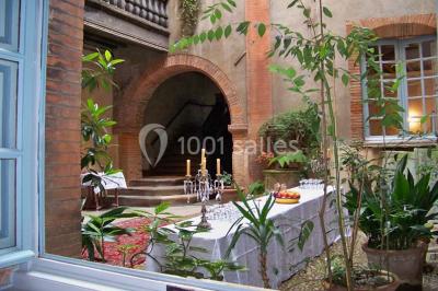 Cour intérieure pavée avec des plantes en pots, entourée de murs en briques et fenêtres à carreaux.