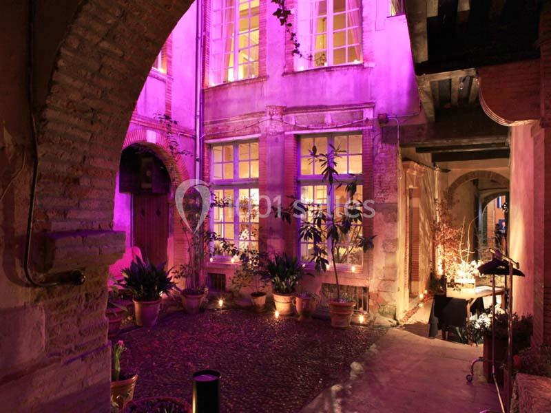 Cour intérieure en soirée, éclairée par des lumières violettes, avec des plantes en pots et des fenêtres illuminées.
