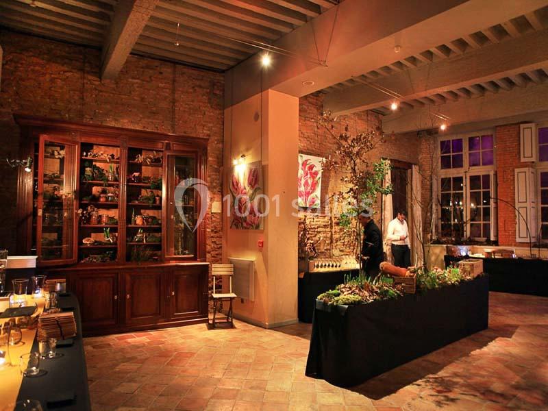 Salle chaleureuse avec murs en briques, poutres apparentes, meubles en bois et tables décorées pour un événement.