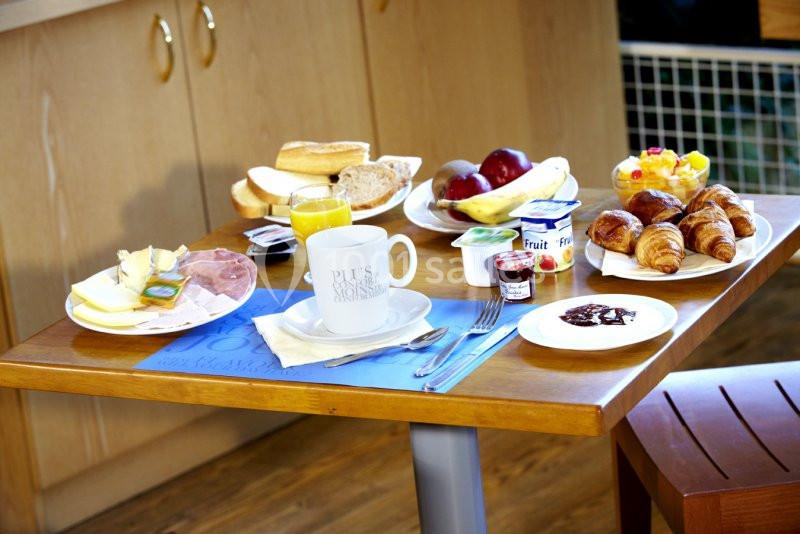 Petit-déjeuner varié sur une table, comprenant viennoiseries, fruits, pain, charcuterie, jus d'orange et yaourts.